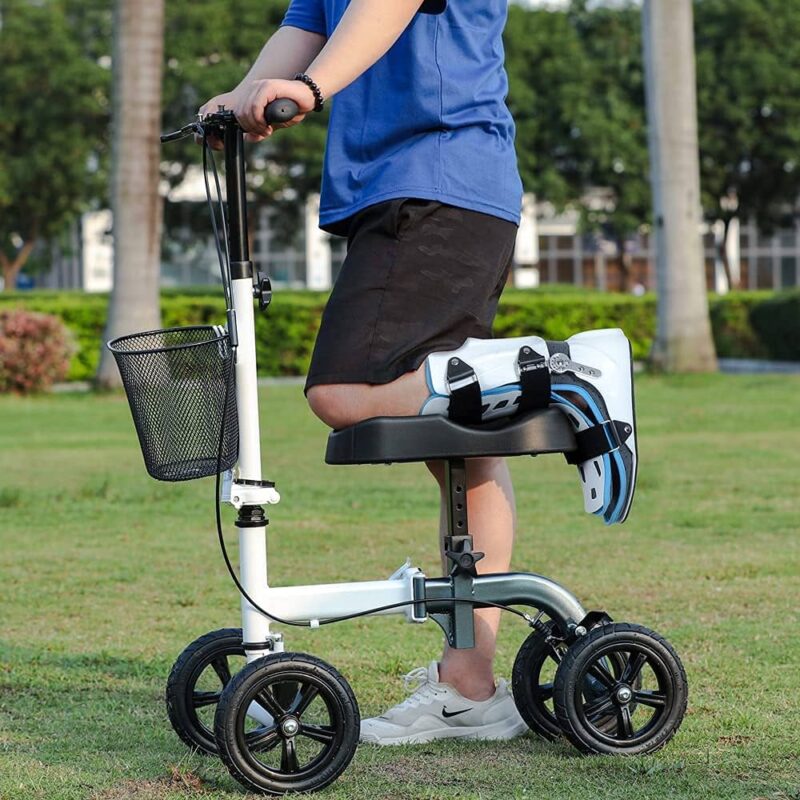 تُظهر الصورة شخصًا في الهواء الطلق، على الأرجح في حديقة أو منطقة عشبية، يستخدم سكوترًا أبيض للركبة. ساقه المصابة مستندة على المنصة المبطنة للسكوتر. يبدو أن الجزء السفلي من الساق المصابة مثبت بدعامة أو جبيرة. يحتوي السكوتر على أربع عجلات، ومقود للتوجيه، وسلة سلكية سوداء صغيرة مثبتة على العمود الأمامي. يستخدم الشخص ساقه الأخرى لدفع السكوتر للأمام على العشب.