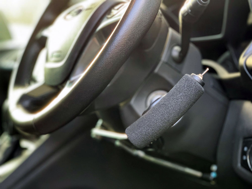 An image of a car's interior shows a specialized driving aid, often referred to as hand controls, installed near the steering wheel. The adaptation consists of a black, textured, cylindrical handle mounted on a metal rod that extends toward the floor, allowing a driver to control the acceleration and braking without using foot pedals. The hand control is positioned to the right of the steering column, and the car's ignition is visible just above the control.